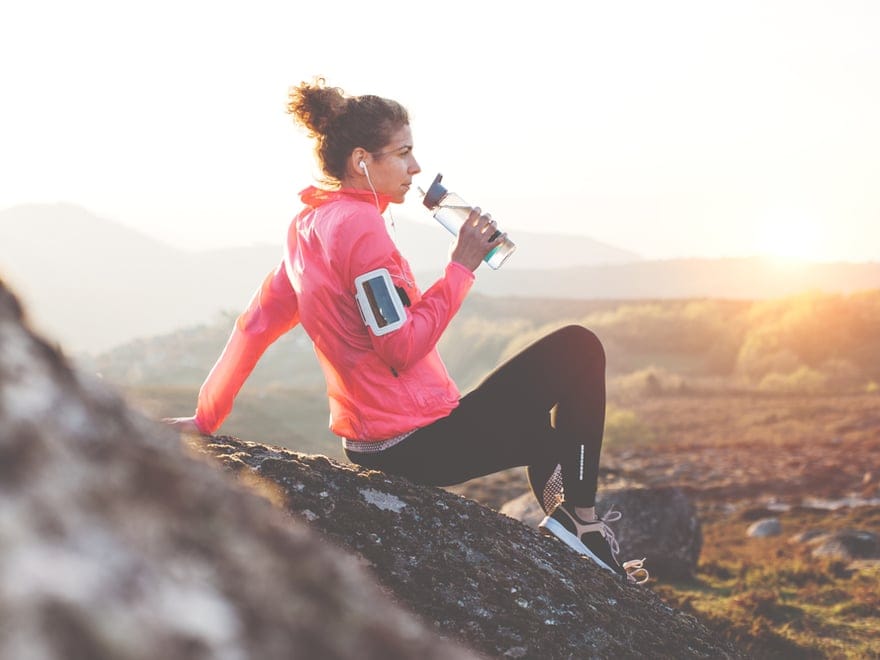 A person in athletic wear and earphones sits on a rocky surface, drinking from a water bottle. They wear a bright pink jacket and black leggings. The background features a sunlit landscape with distant hills and a warm glow. A person in athletic wear and earphones sits on a rocky surface, drinking from a water bottle. They wear a bright pink jacket and black leggings. The background features a sunlit landscape with distant hills and a warm glow.