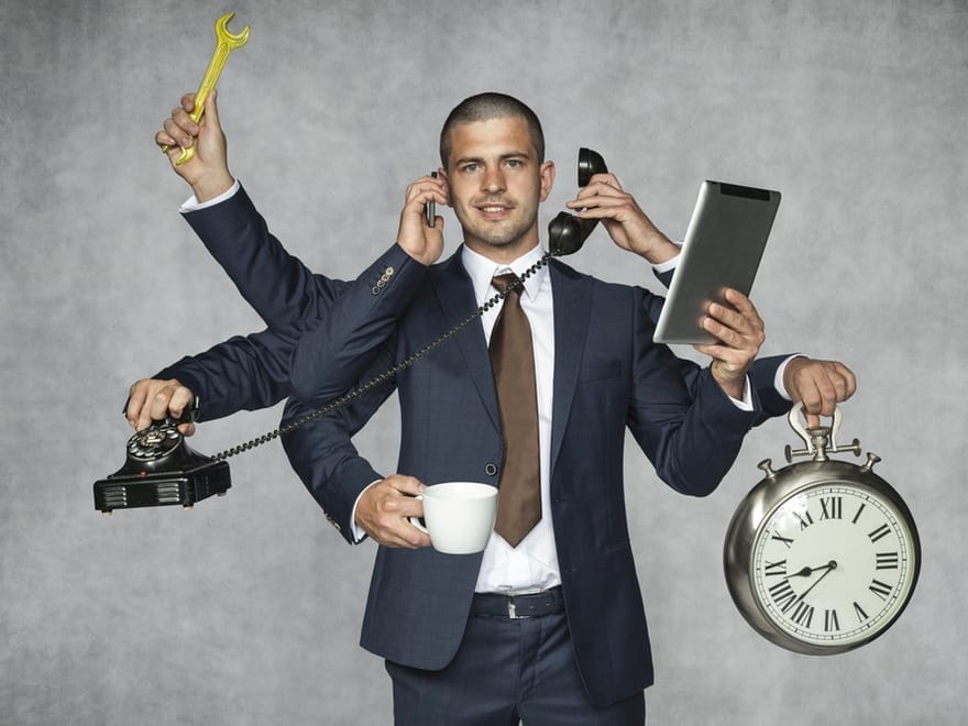 Man in a suit with multiple arms holds various items including a wrench, phone, tablet, coffee cup, and clock, symbolizing multitasking and time management, against a gray background. Man in a suit with multiple arms holds various items including a wrench, phone, tablet, coffee cup, and clock, symbolizing multitasking and time management, against a gray background.